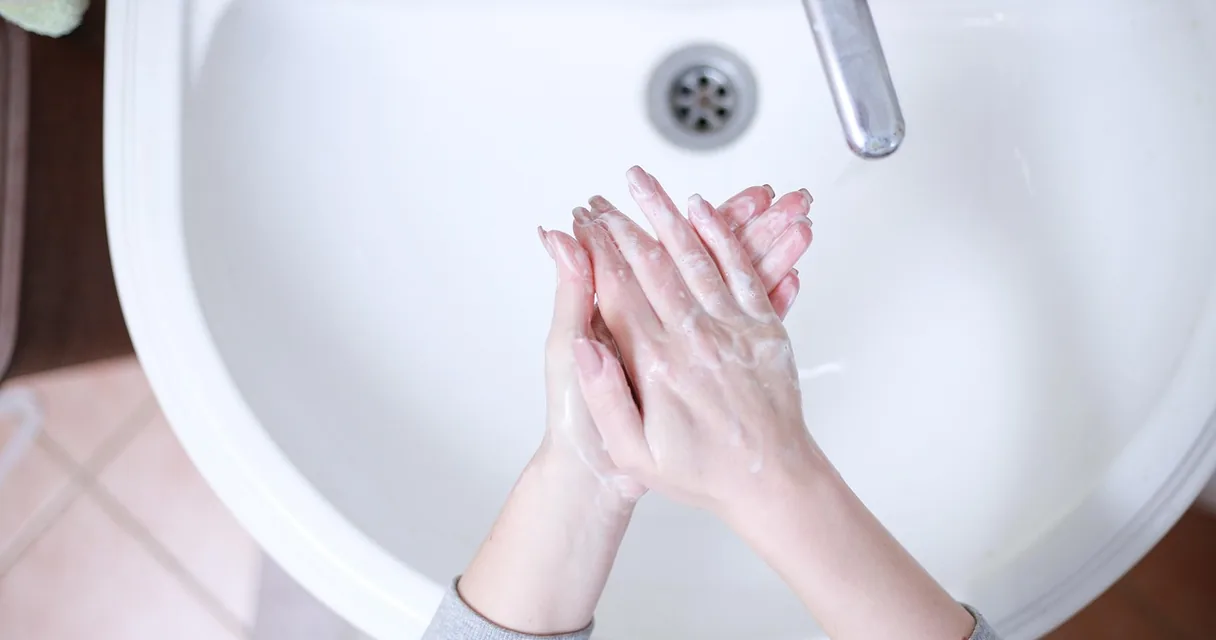 Mãos esfregando espuma de sabonete sobre uma pia branca, ilustrando a pele do rosto caindo com a idade